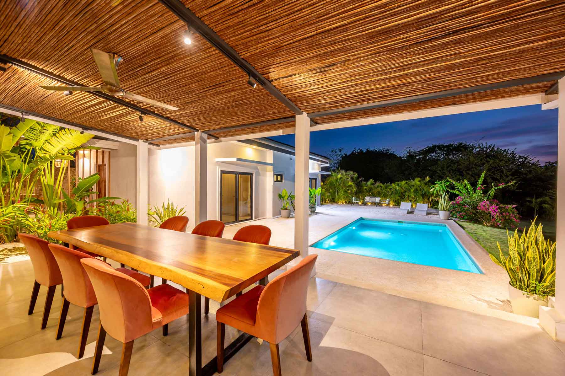 The covered terrace at dusk, with a live-edge dining table for ten beside the lit pool